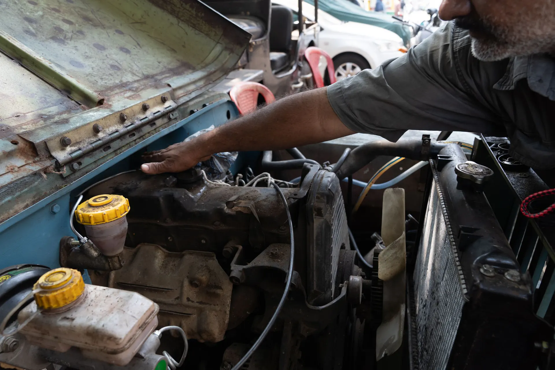 A mechanic reaches into the engine bay of an older jeep at an open-air workshop.