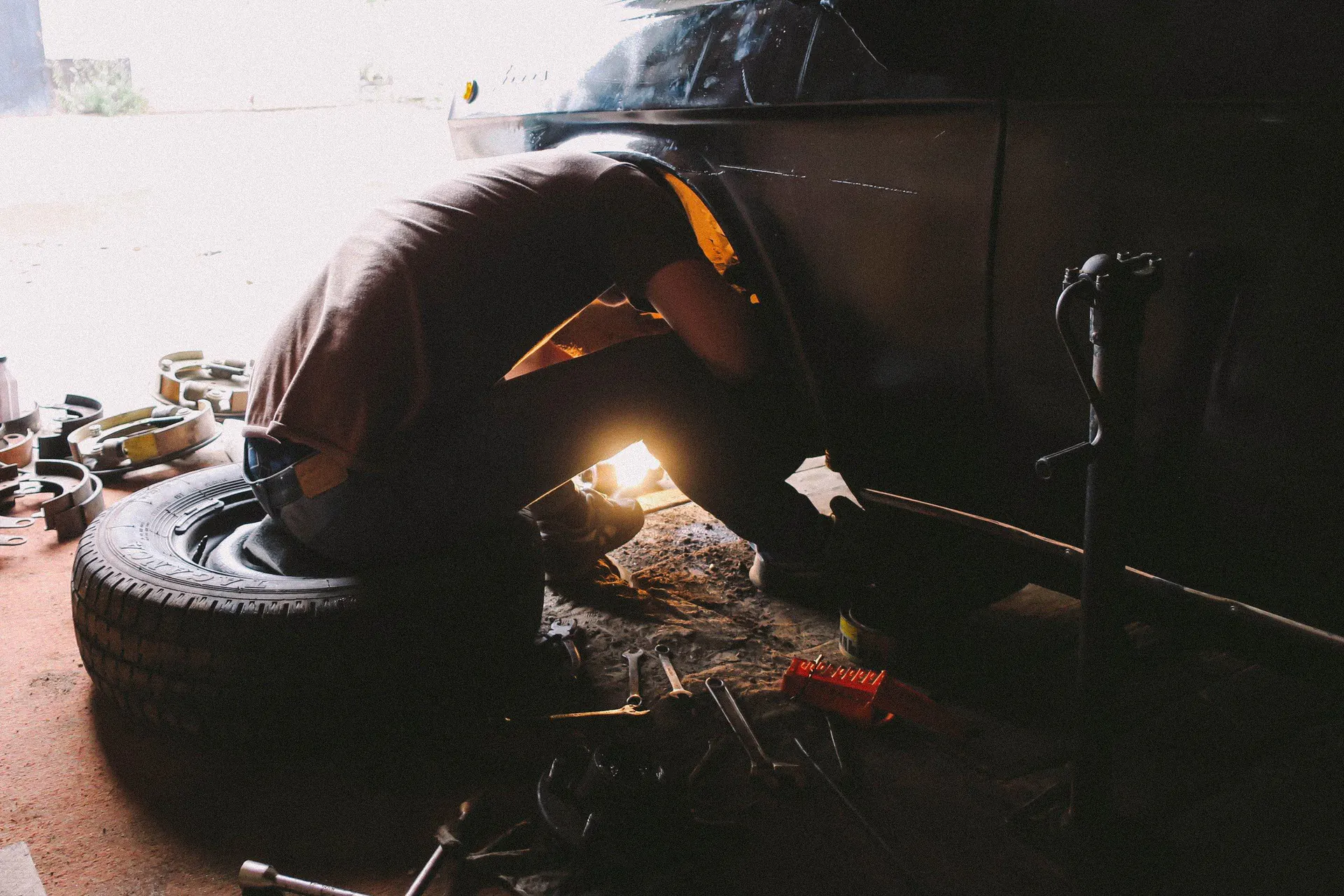 A mechanic crouches by a car wheel in a dim workshop, lit by a hand-held work light.